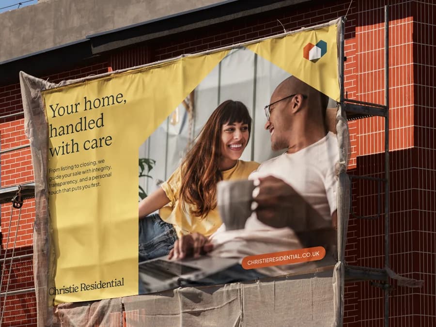 A yellow Christie Residential banner lashed to scaffolding on a brick building, the brand line and mark still reading clearly at distance and half in shadow.