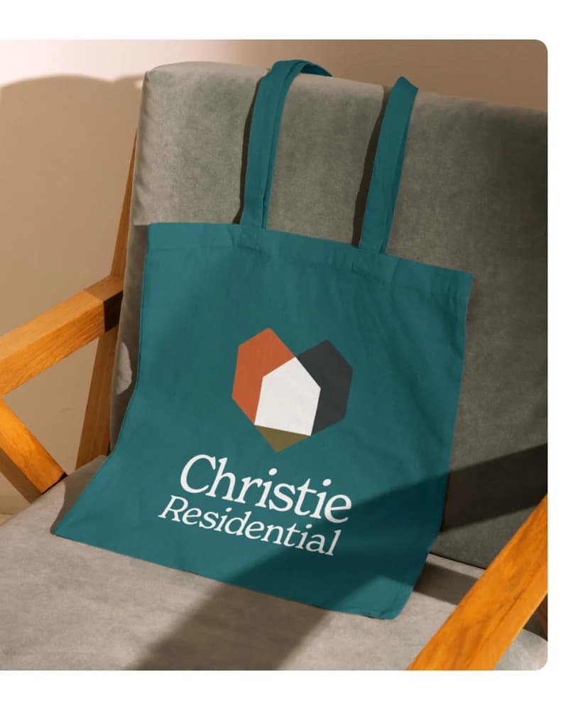 A teal Christie Residential tote bag hanging on a wooden chair, the house mark and wordmark printed in white.