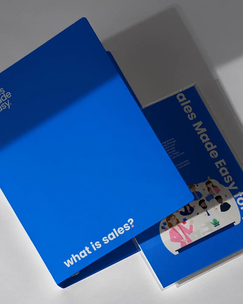 Sales Made Easy printed folders and a brochure fanned on a pale surface, cobalt on navy, the cover asking 'what is sales?'.